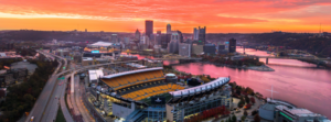 Pittsburgh skyline and Acrisure Stadium glowing at sunset, with the Three Rivers and city landscape creating a stunning aerial view of the Steel City.