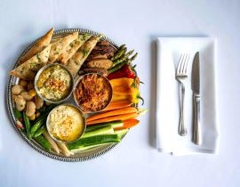 three hummus dishes on a platter surrounded with veggies, began sausage and pita bread